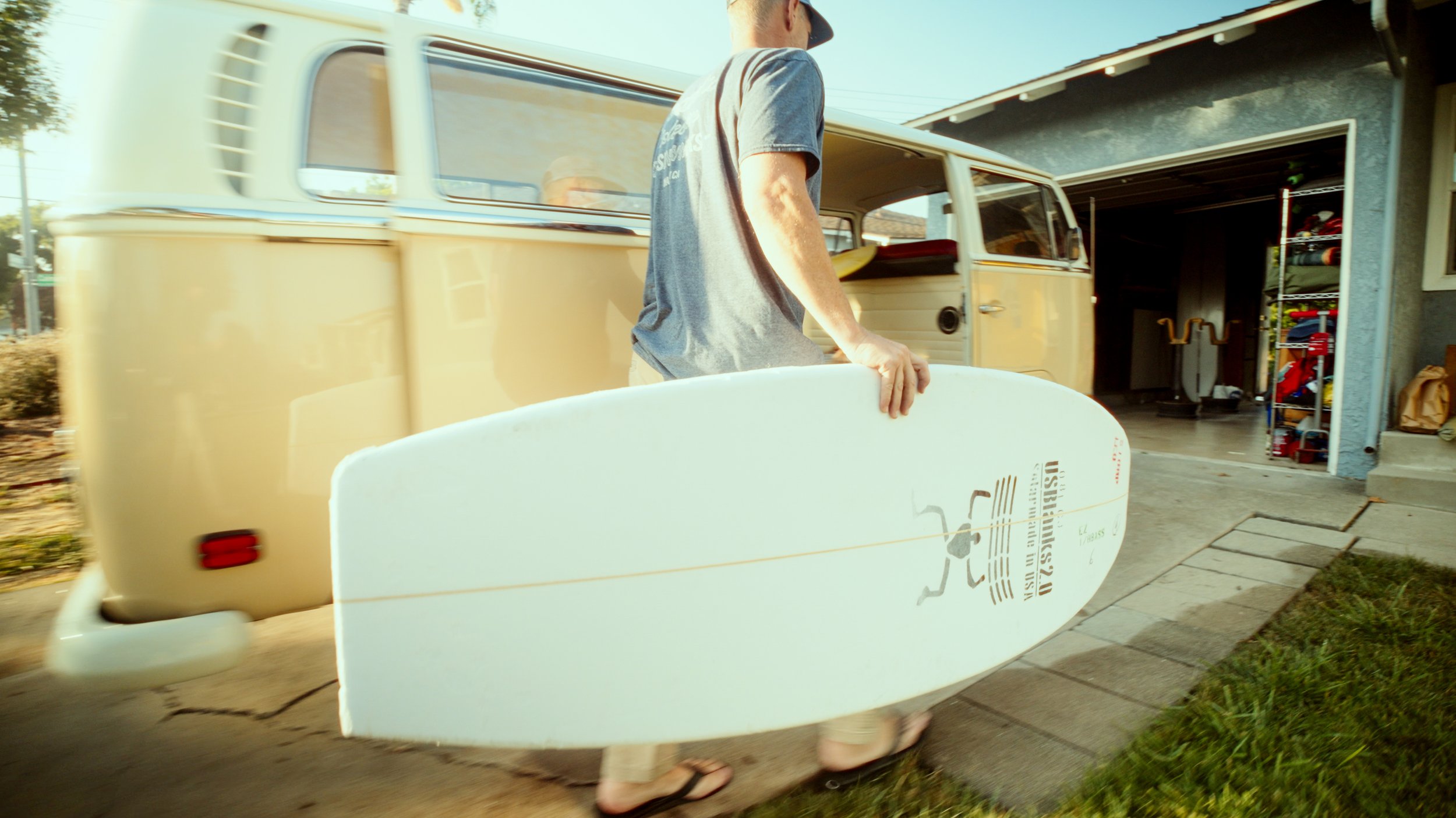 A person holding a white surfboard walking towards a yellow vintage van parked outside a garage.