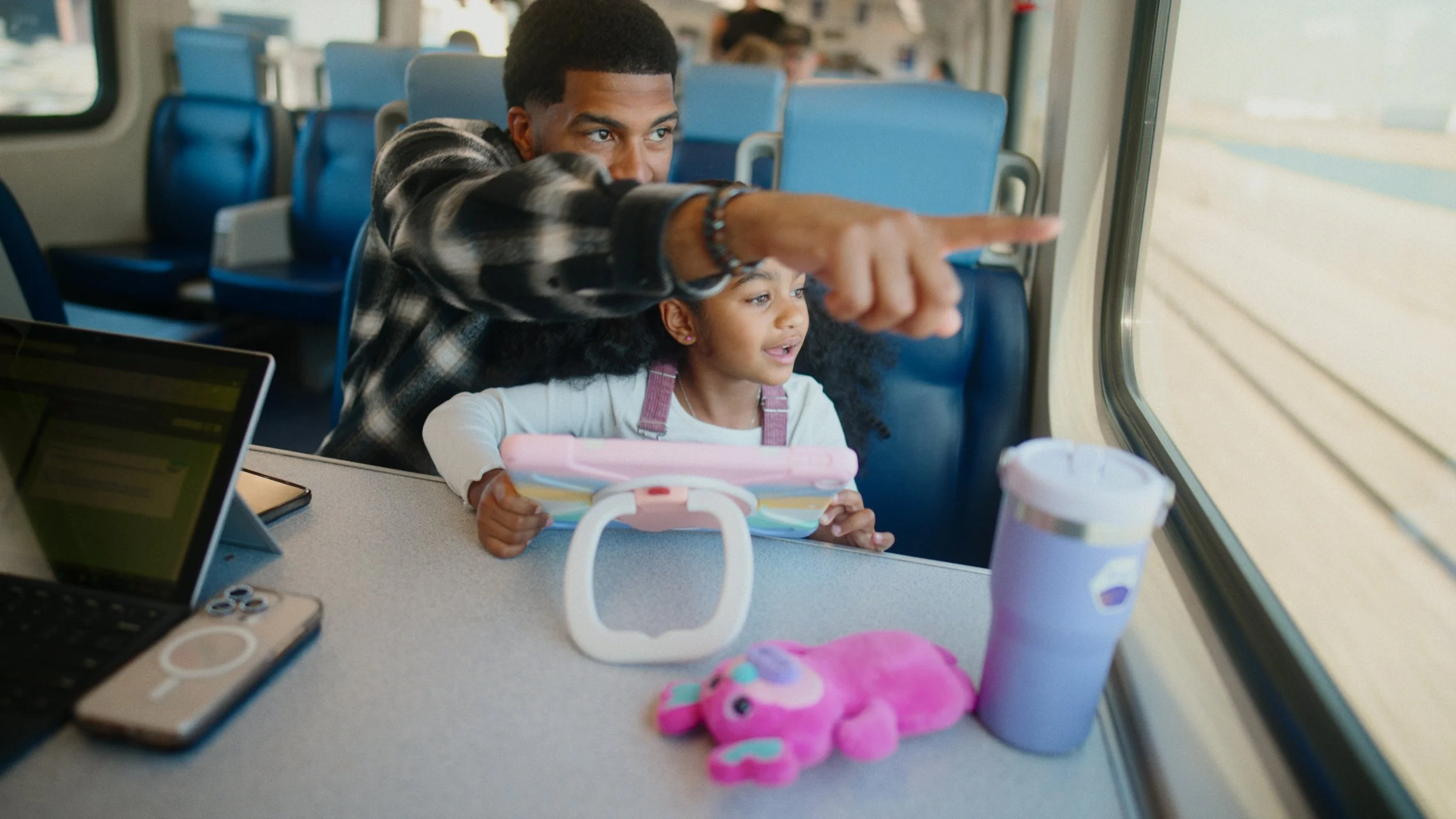 A young girl and a man, possibly her father, sitting together on a train and looking out the window. The girl is holding a tablet, and there are toys, a water bottle, and a laptop on the table in front of them.