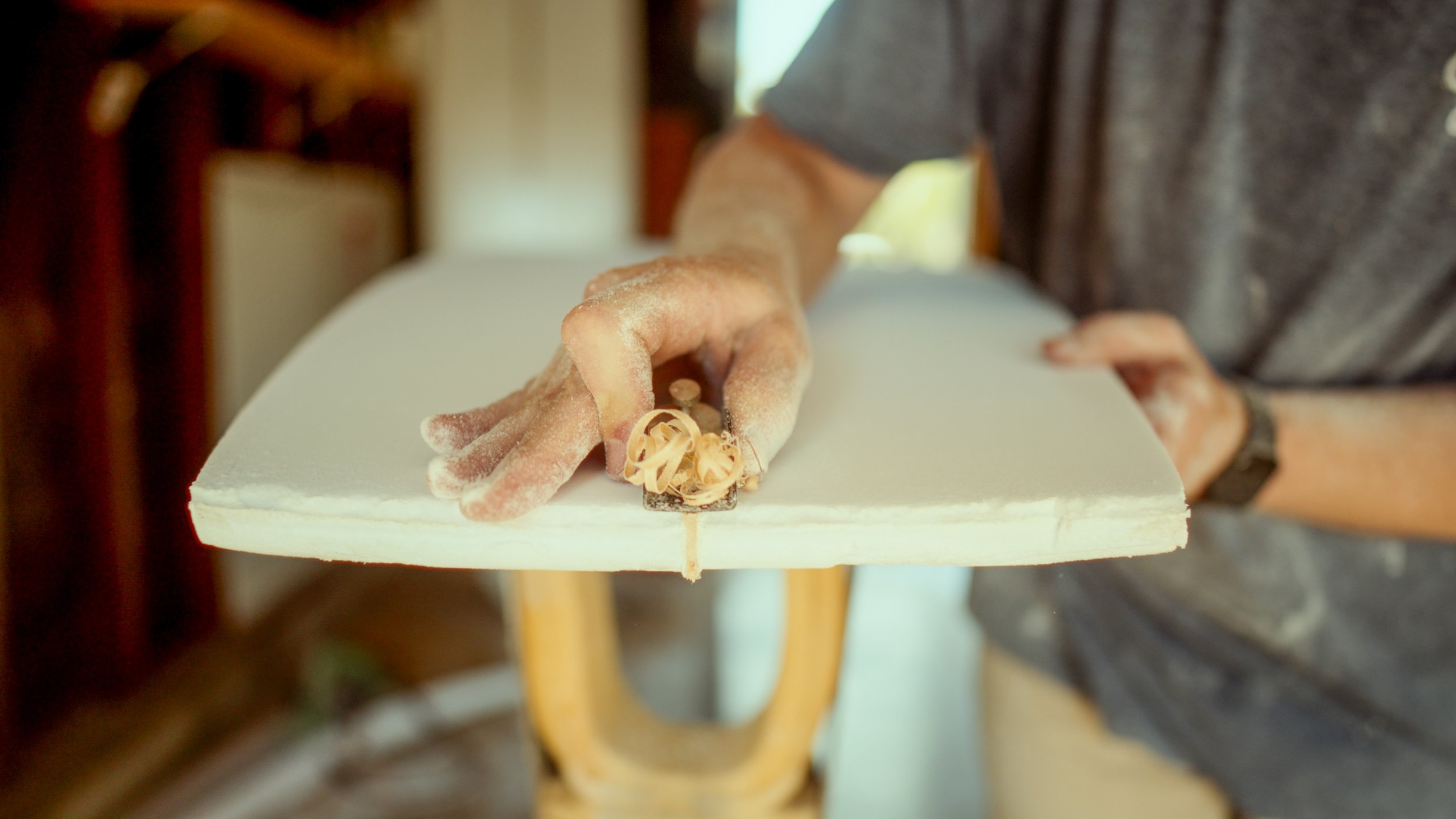 Person sanding a wooden surfboard with a handheld sander, with wood shavings on the surface.