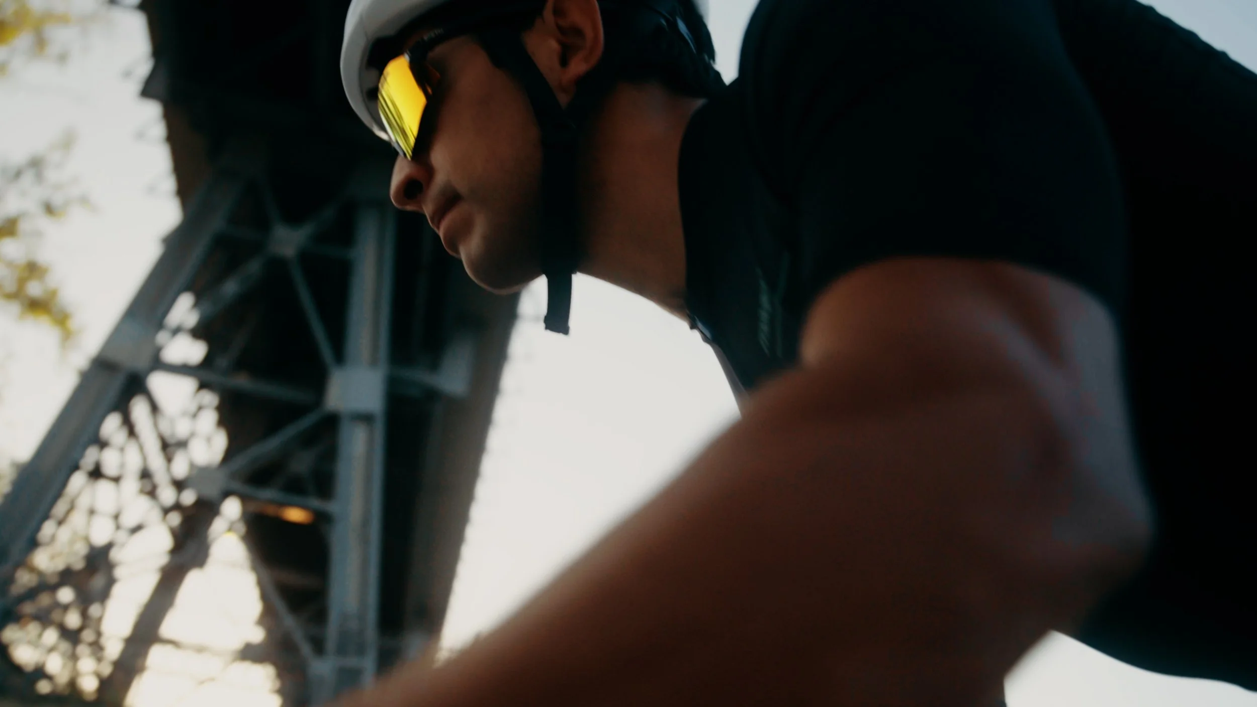 A cyclist wearing a helmet and sunglasses, captured from a low angle, with a metal bridge structure in the background.