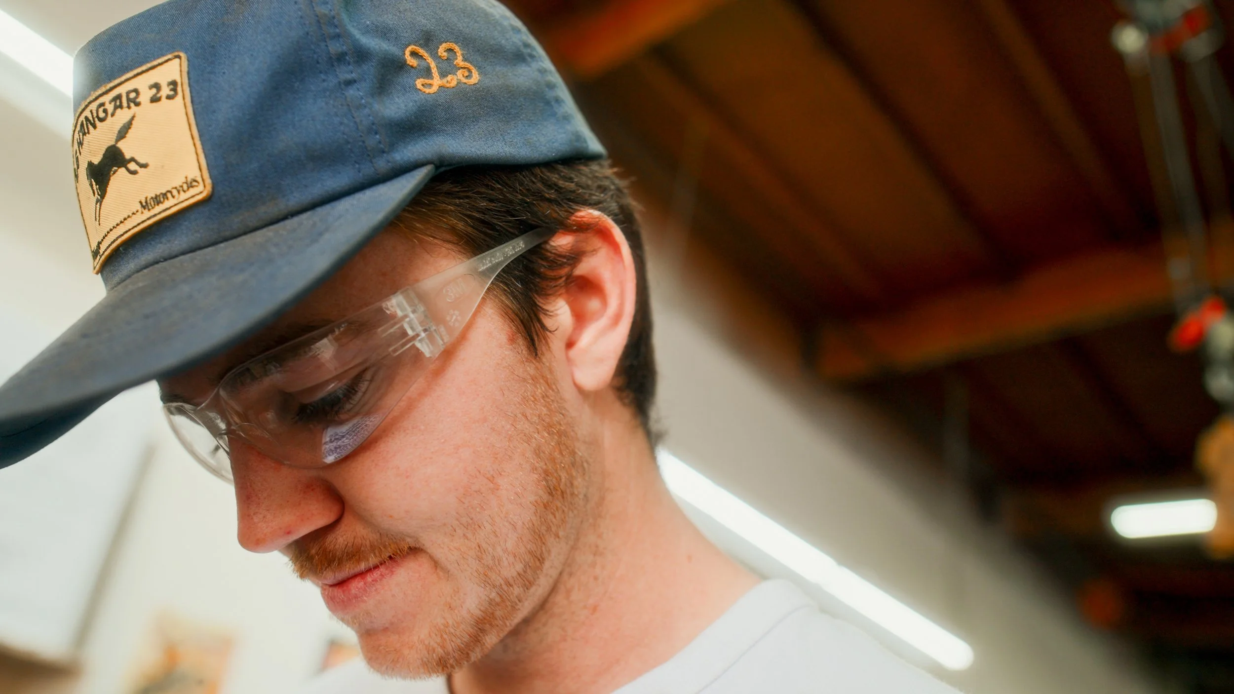 Close-up of a young man wearing a blue cap with a patch that says 'BUNGAR 23' and a logo of a kangaroo, and transparent protective glasses, looking down.