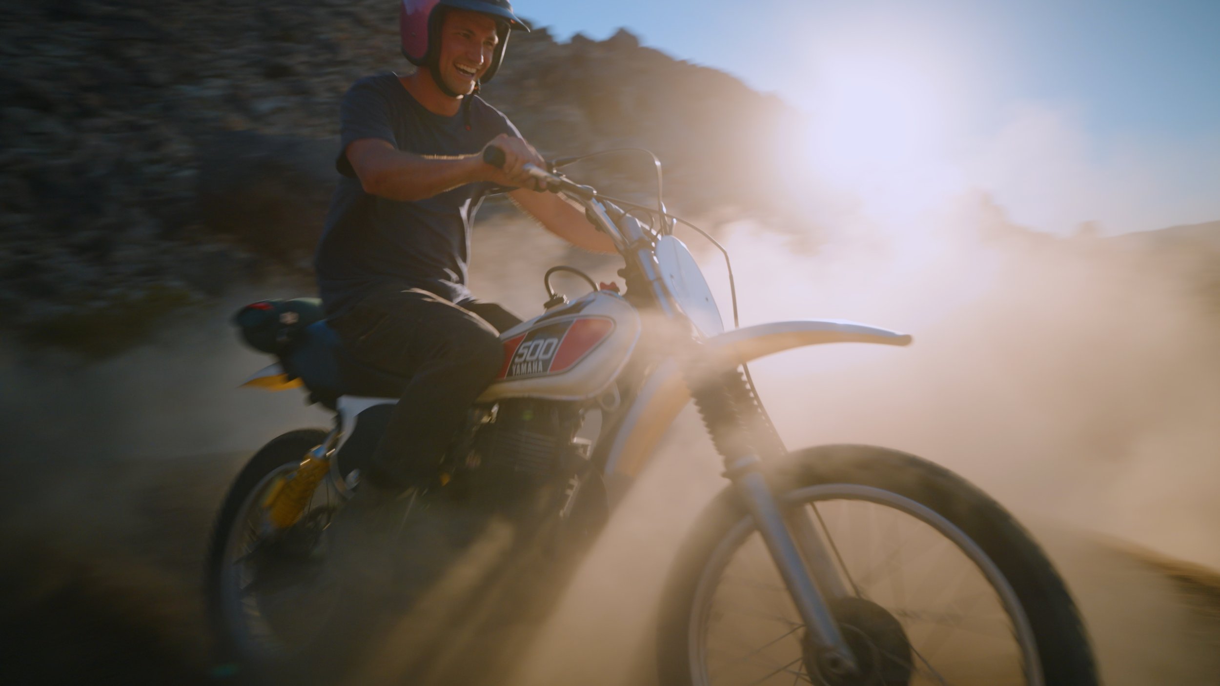 A man riding a motorcycle through a dusty dirt trail with the sun low in the sky.