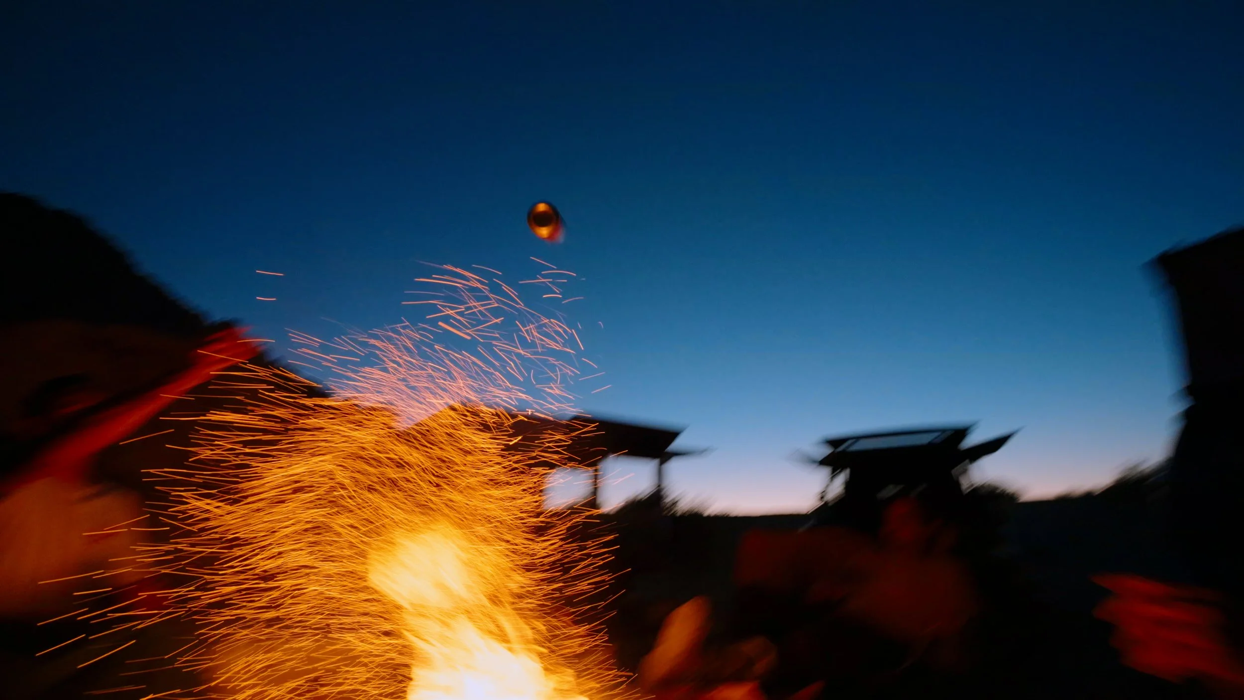 People around a bonfire at dusk with sparks flying into the air, and a ball flying in the sky.