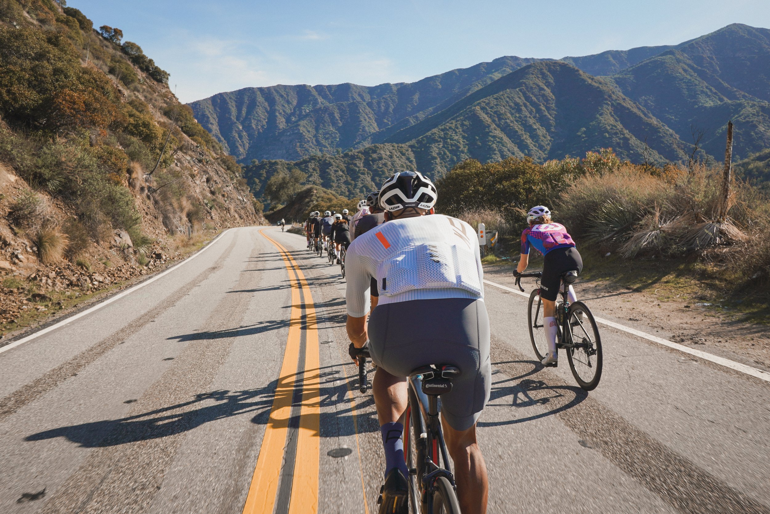 Cyclists riding on a mountainous road with hills and trees in the background on a sunny day.