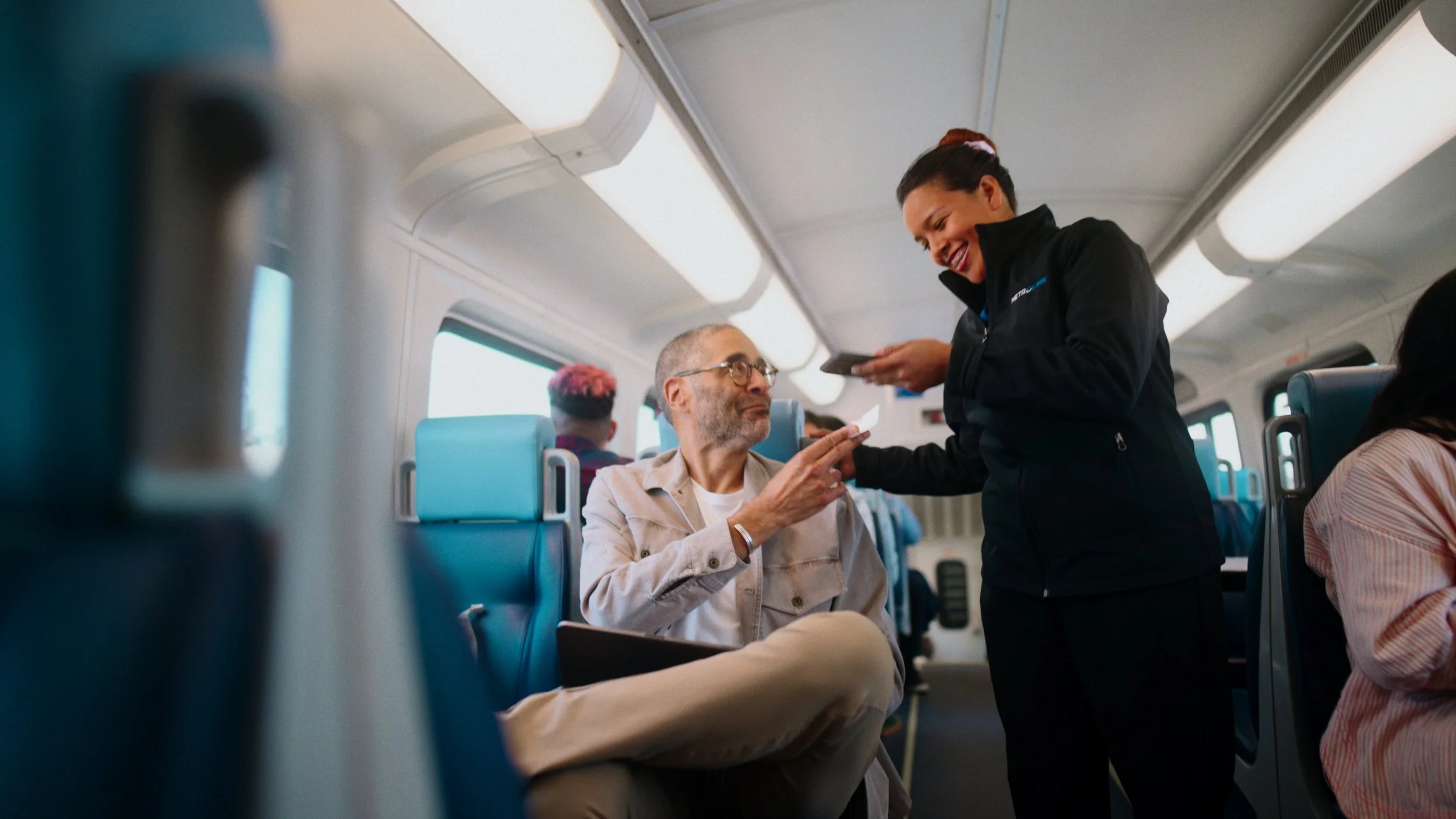 A happy woman in a black jacket is handing a boarding pass or ticket to a seated man with glasses on an airplane, with other passengers in the background.