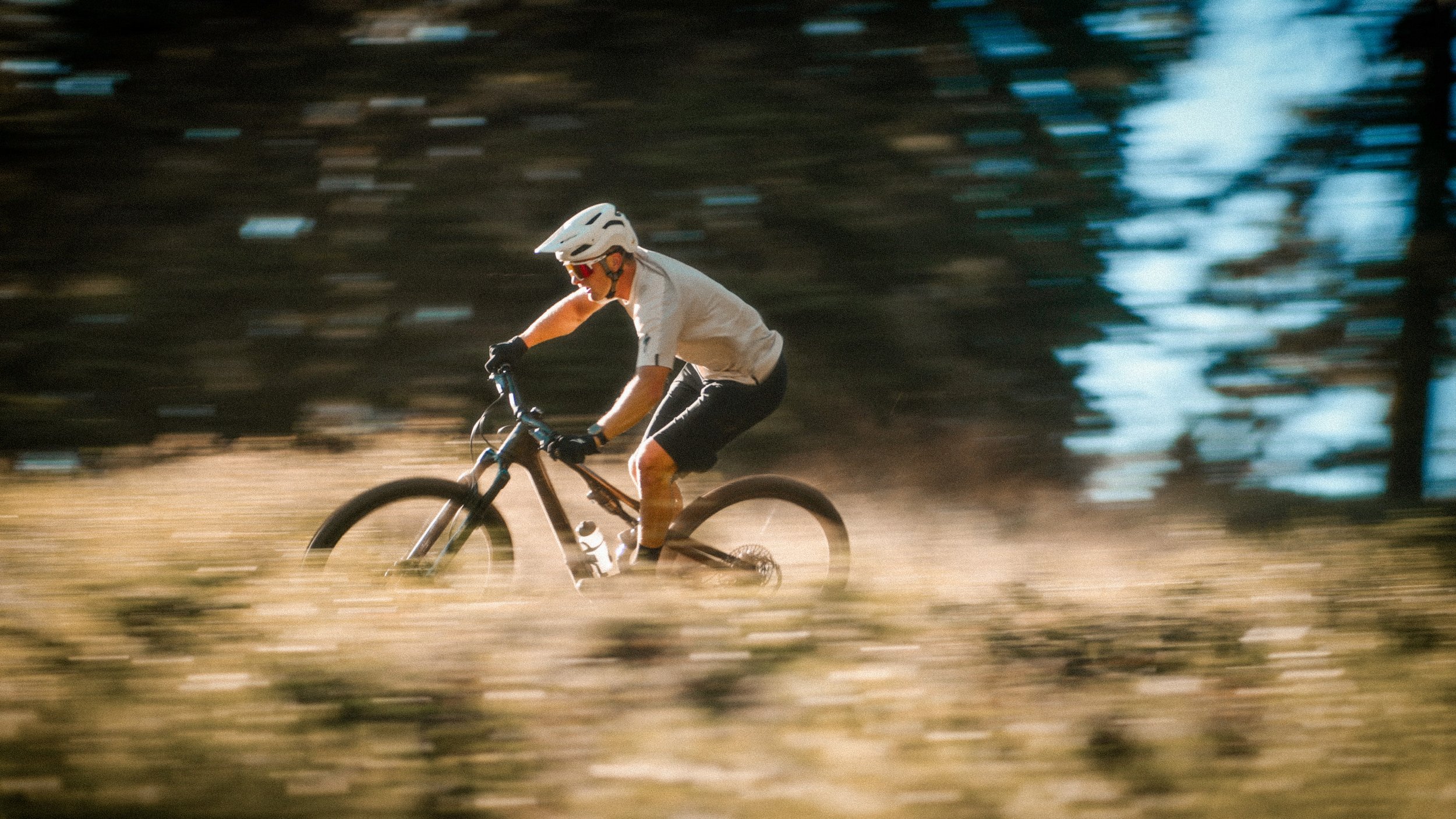 A person riding a mountain bike through a blurred outdoor trail, wearing a helmet, sunglasses, a white shirt, and black shorts.