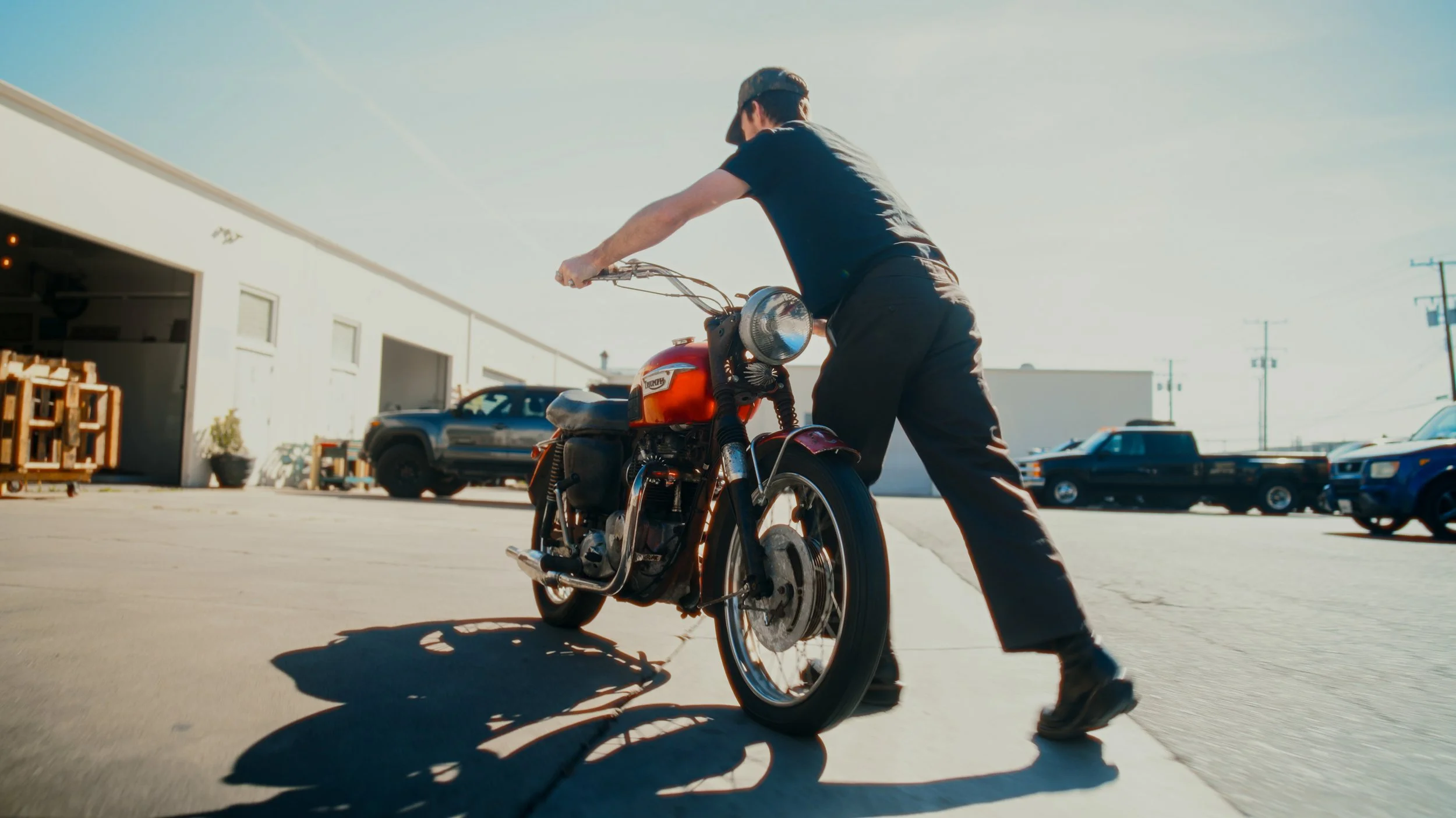 Person pushing a vintage Triumph motorcycle in an outdoor parking lot with parked cars and a warehouse in the background.