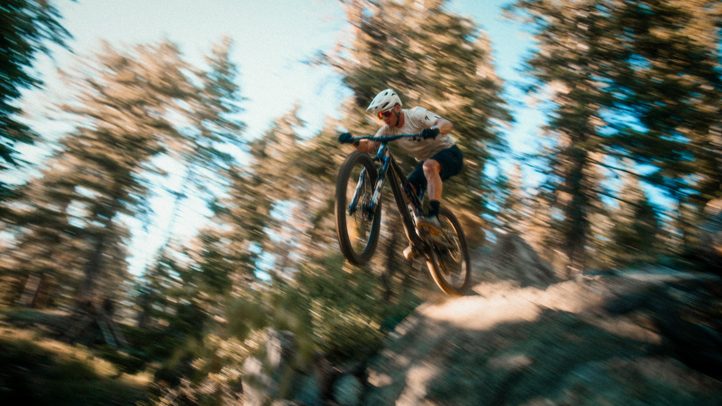 Person wearing helmet and sunglasses riding mountain bike off a jump on a dirt trail through a forest at sunset.