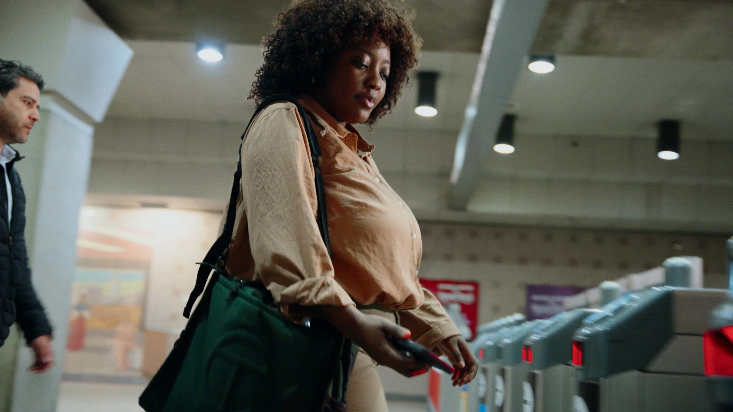 A woman with curly hair wearing a beige shirt and carrying a black bag is holding a phone and standing near a ticket gate in a public transportation station. A man is visible in the background.