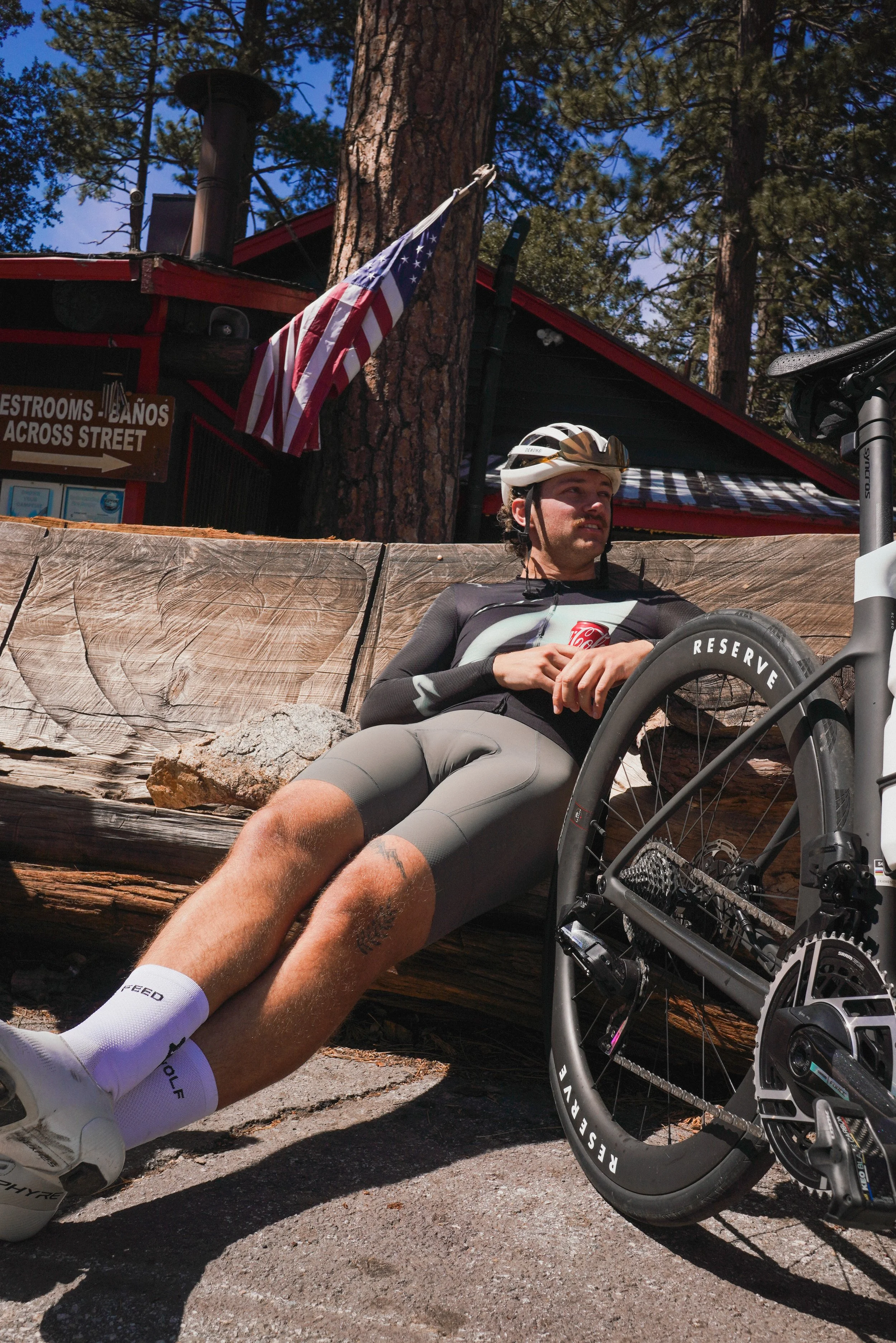 Cyclist resting on a log next to his bike, holding a soda can, wearing a helmet and cycling outfit, with an American flag and a building in the background.