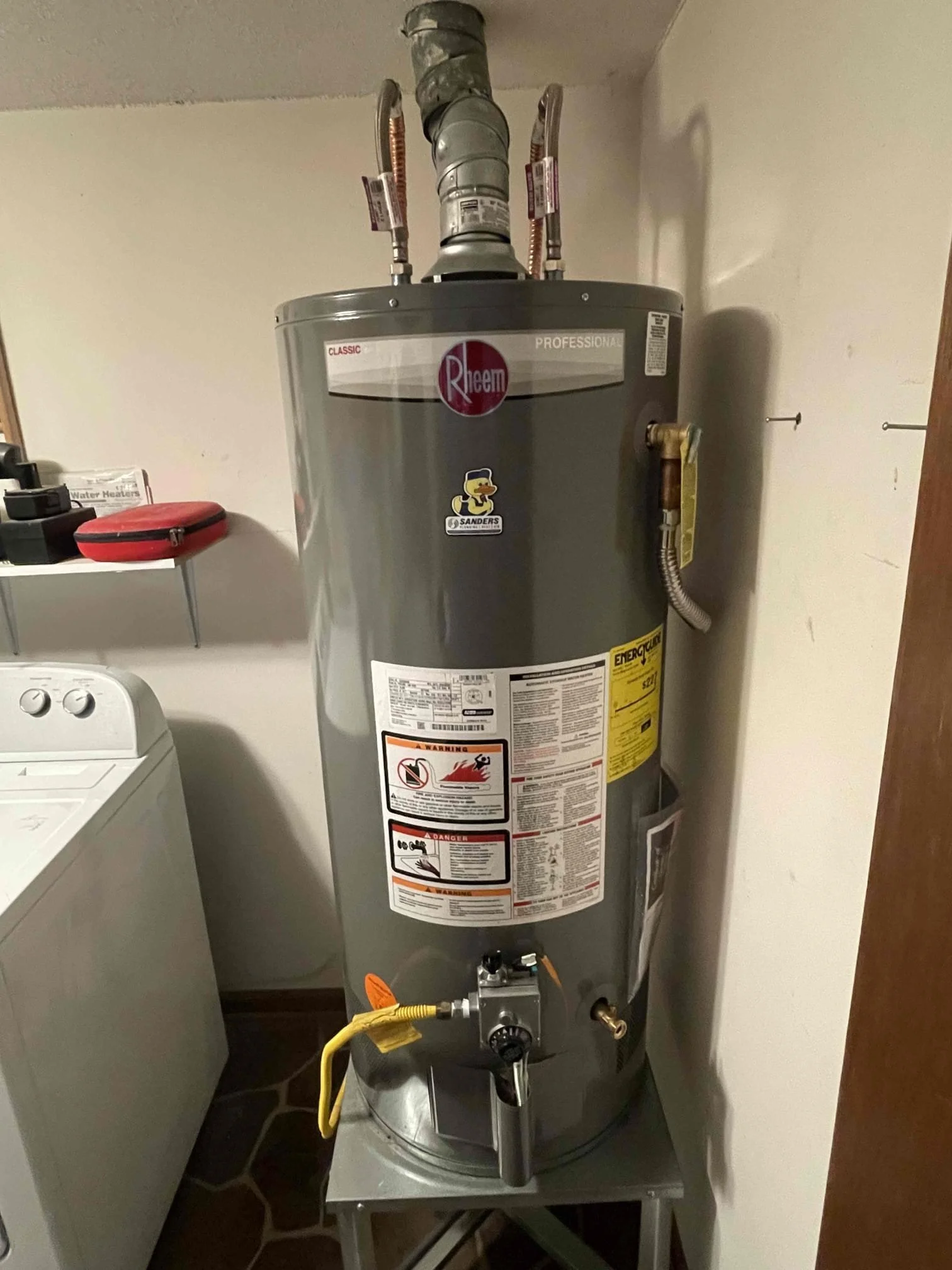 A gray Rheem water heater in a laundry room, positioned next to a white washing machine. The water heater has various warning stickers, a brass drain valve at the bottom, and copper pipes connected at the top.
