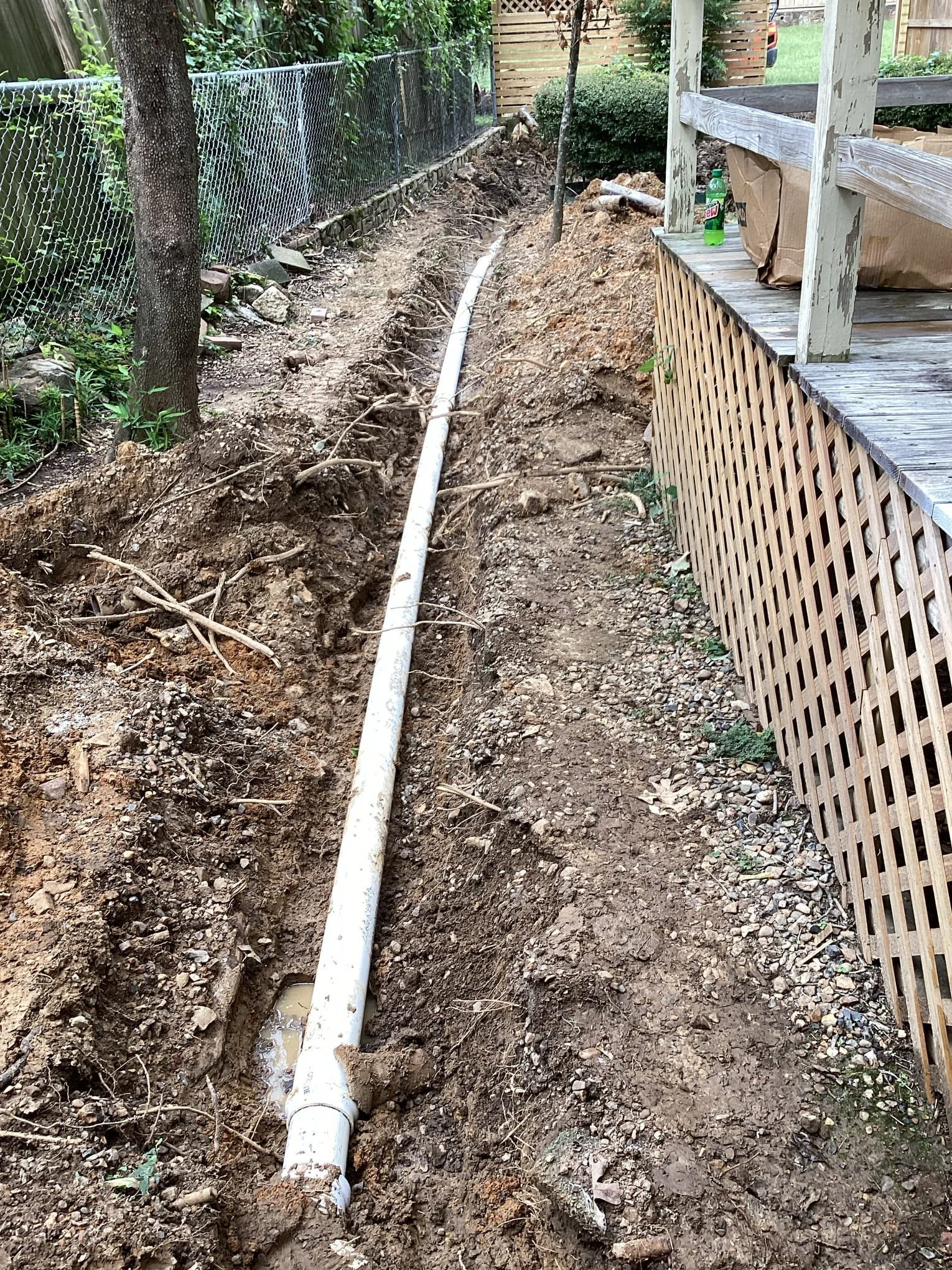 A long trench dug along a backyard fence with a white PVC pipe laid inside. The trench has exposed soil and roots, with a wooden deck on the right and a chain-link fence on the left, surrounded by greenery.