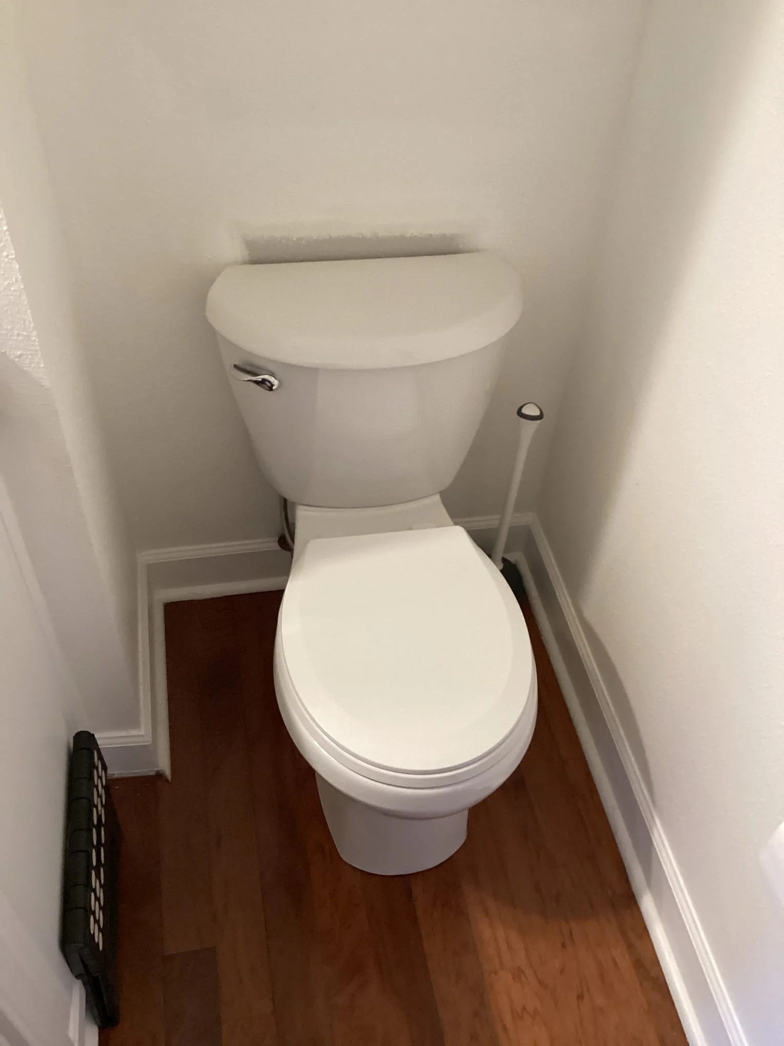 A standard white toilet in a small bathroom with wooden flooring, located between two white walls.