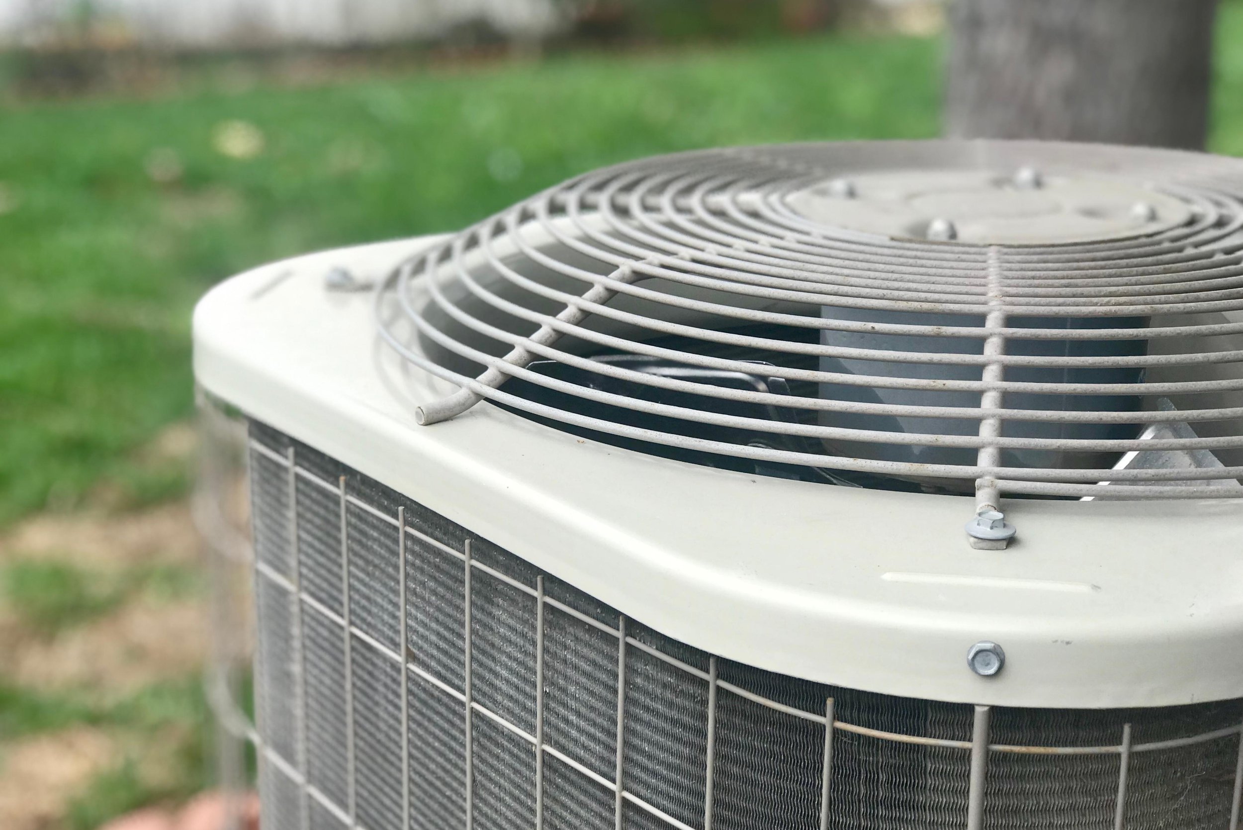 Close-up view of an outdoor air conditioning unit with a metal protective grille on top, situated in a grassy area with a tree in the background.