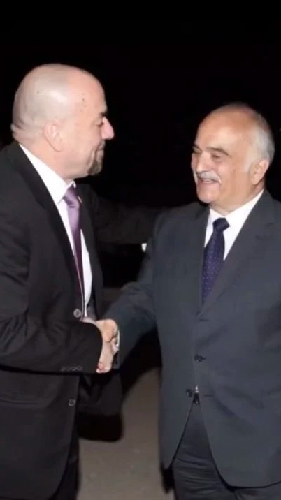 Two men in suits shaking hands and smiling at each other outdoors at night.