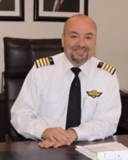 Man in airline pilot uniform sitting at desk, smiling, with framed photos on the wall behind him.