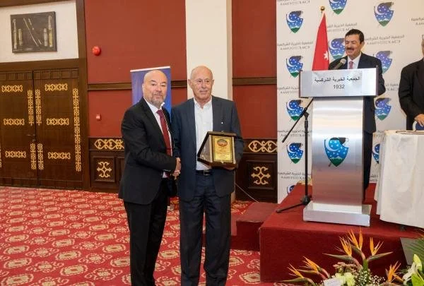 Two men shaking hands, one holding a plaque, in a formal setting with a podium and a backdrop with logos and text in Arabic. A third man stands at the podium.