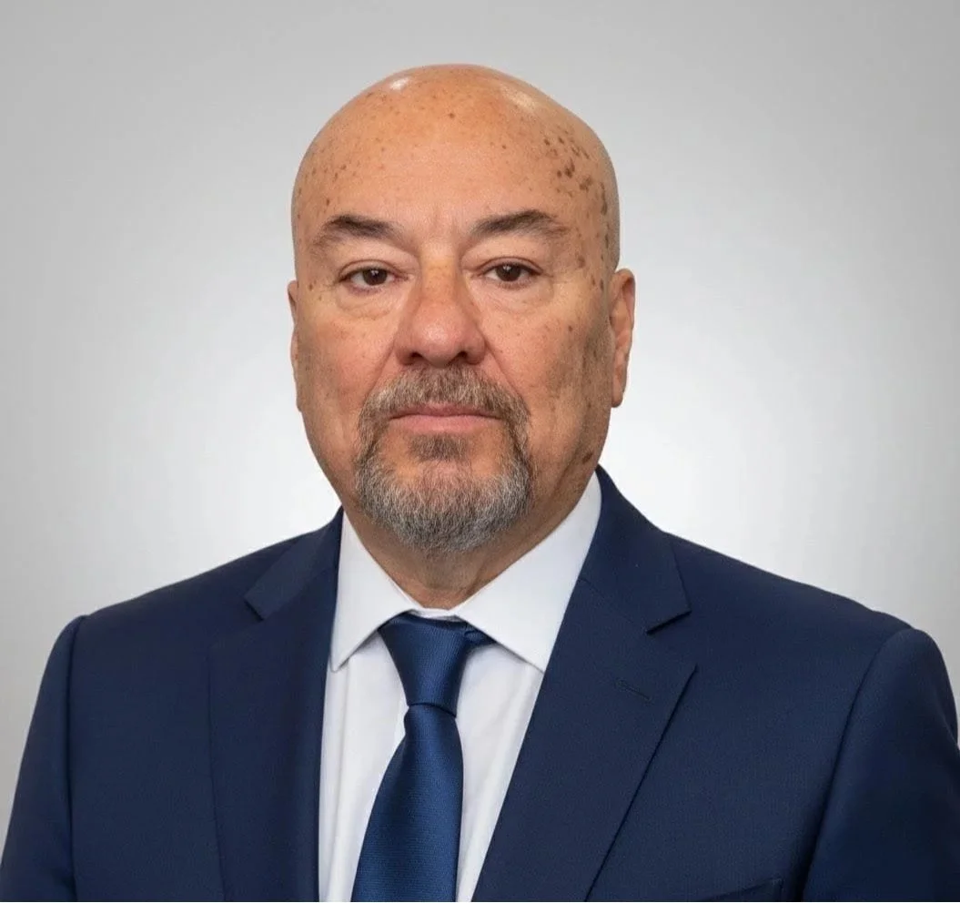 Headshot of a bald man with a goatee, wearing a dark suit, white shirt, and blue tie, against a plain gray background.