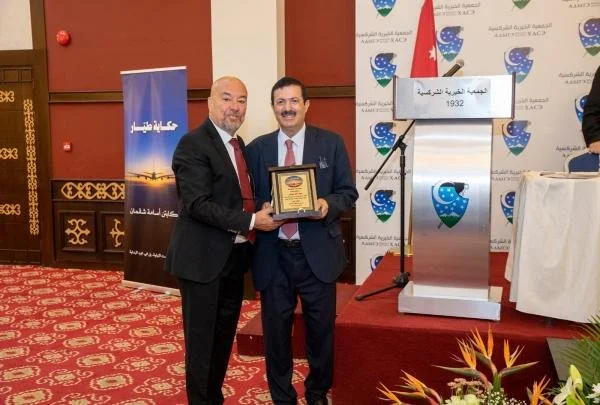 Two men in suits, one holding a plaque, standing on a stage with a backdrop displaying a logo and text in a foreign language. A lectern and floral arrangement are visible.