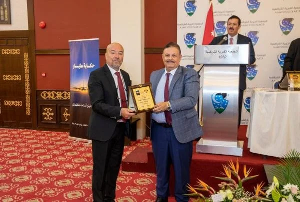 Two men in suits are standing and smiling at a ceremony, one of them holding a framed award or certificate. There is a speaker at a podium behind them and flags in the background. The setting appears formal with a patterned red carpet and decorative 