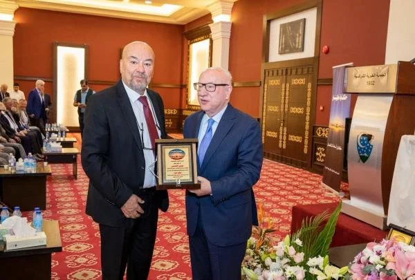 Two men in suits standing in a decorated banquet hall. One man is handing a plaque to the other. There are people seated at tables in the background and floral arrangements in the foreground.