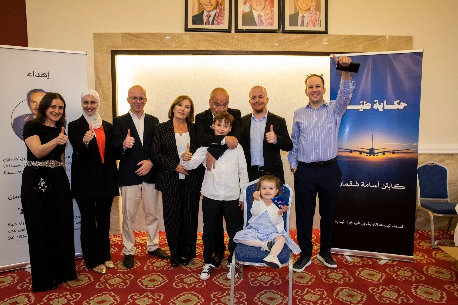 A group of ten people, including two children, posing together at a formal event, with some giving thumbs up and smiling. Behind them are banners with Arabic writing and an airplane image, and the setting appears to be a conference or banquet room wi