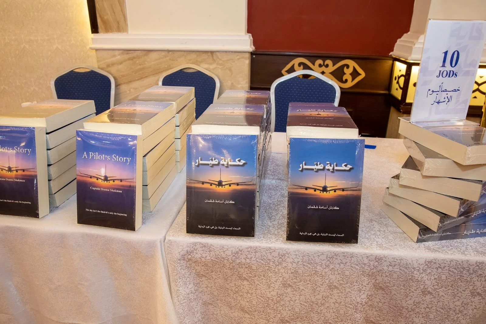 Books titled 'A Pilot's Story' and in Arabic, displayed on a table at a book event, with a sign indicating a price of 10 JODs.
