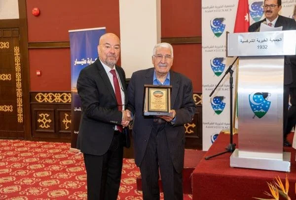 Two men in suits shaking hands; the man on the right holds a framed award or plaque; background shows a speaker at a podium and banners with Arabic and crescent moon and star symbols.