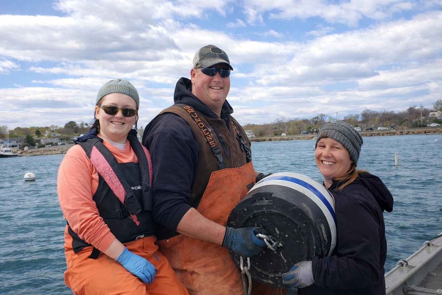 Three people smiling, wearing outdoor clothing and gloves, holding a funnel with a chain, standing near water with houses and trees in the background.