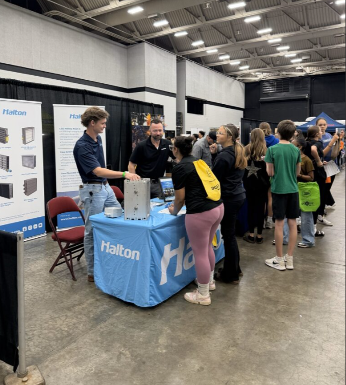 People attending a trade show or conference, gathered around a booth with a blue tablecloth featuring the 'Halton' logo, engaging with representatives and learning about industrial equipment or technology.