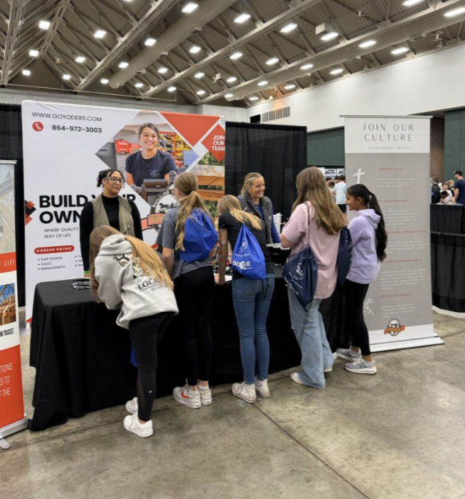 Group of young women at a career fair booth with signage promoting construction careers and a company website, inside a large convention center.