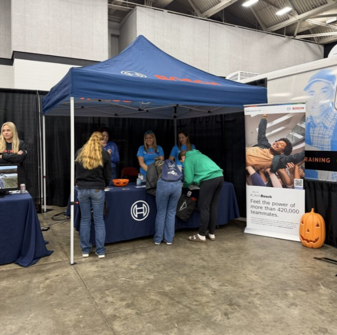 People gathered under a blue canopy tent at an indoor event. There are four women behind the table wearing blue shirts, and a few people in front of the table engaging with them. A promotional banner with a person lying down and the Bosch logo is nea