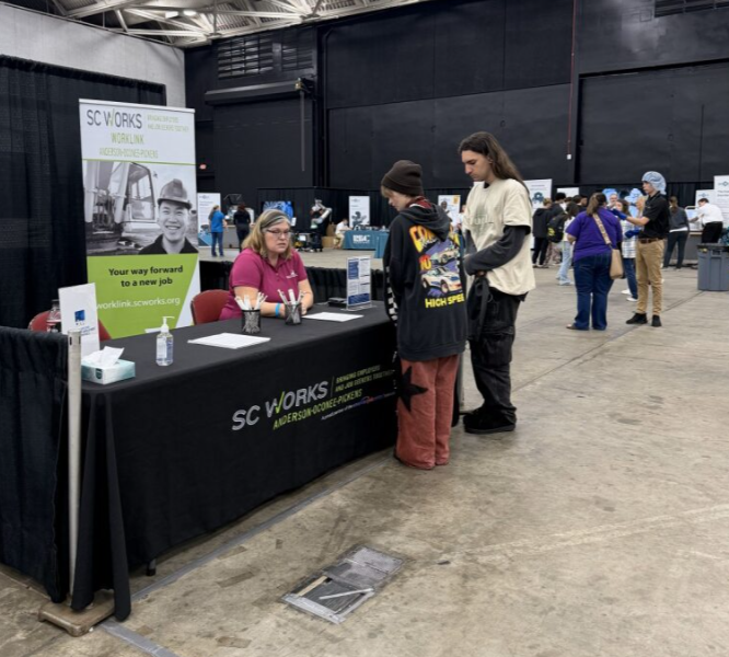 People at a career fair booth for SC Works, with a woman sitting at a table talking to two visitors. Other booths and attendees visible in the background.