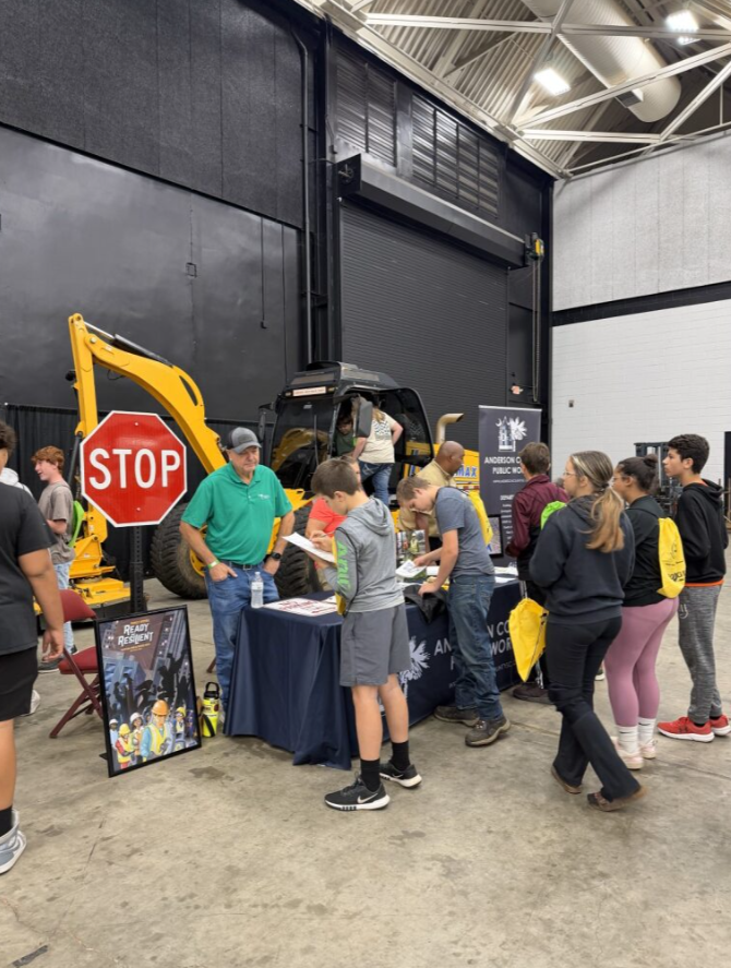 Group of people gathered around an informational booth at an indoor event with heavy machinery and construction-themed displays.