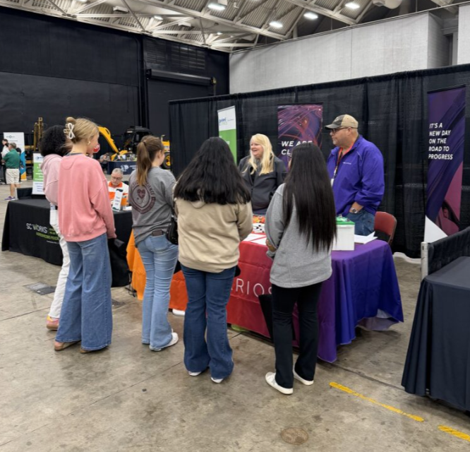 Group of five women and two men talking at a booth in a conference or trade show exhibition.