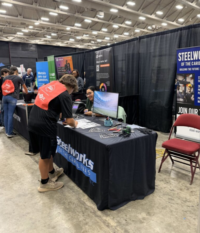 At a trade show or career fair booth for Steelworks of the Carolinas, a person in a black shirt and shorts is signing a document while engaging with a woman at the table. The booth features a monitor, flyers, and banners promoting Steelworks and care