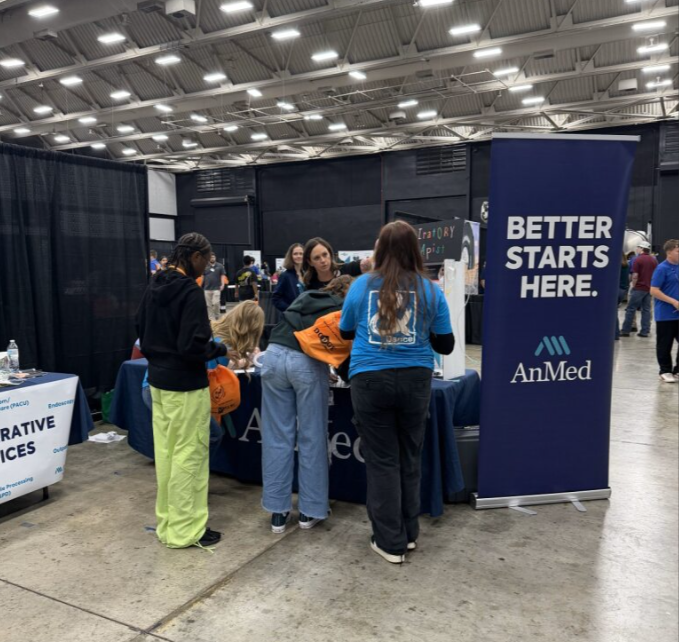 People gathered around an informational booth at a convention or expo, with a tall blue banner that reads 'Better Starts Here' and features the AnMed logo, inside a large indoor event space with high ceilings.