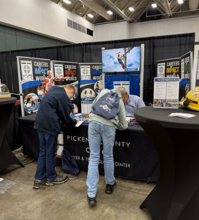 People at a career and technology center booth at an event, with one person signing a paper, another person with a backpack, and a man behind the table, with a screen showing a worker on a power line in the background.
