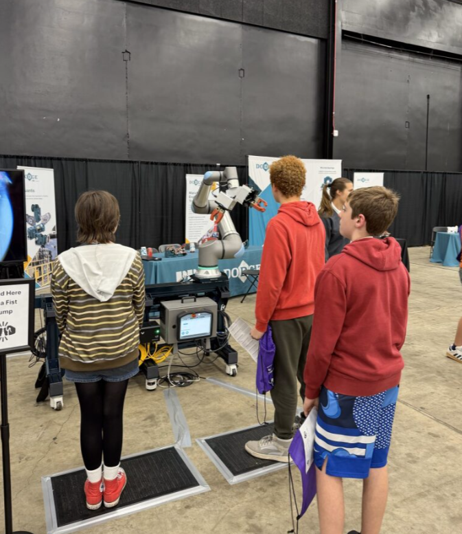 Kids observing a robotic arm demonstration at an engineering event, with tables and display boards in the background.