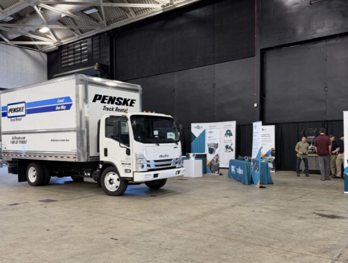 A Penske truck is displayed inside a large industrial building or convention center with a few people and promotional stands nearby.
