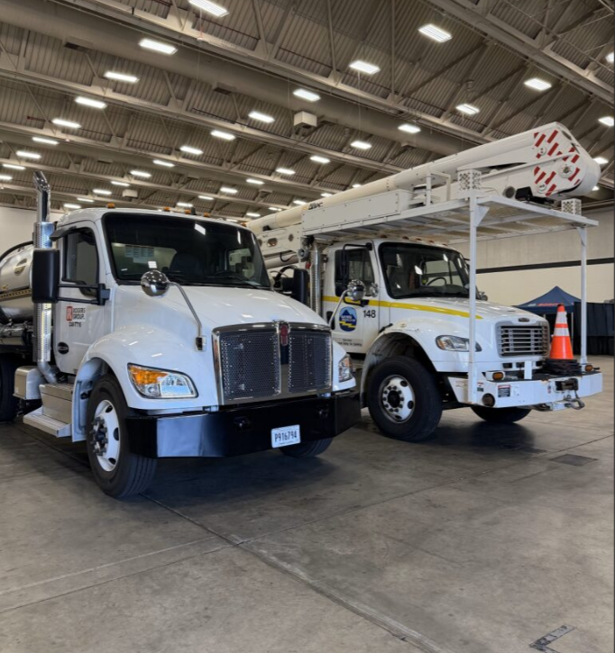 Two white utility trucks, one with a large rocket or missile on a lift and a traffic cone, parked inside a spacious indoor facility.