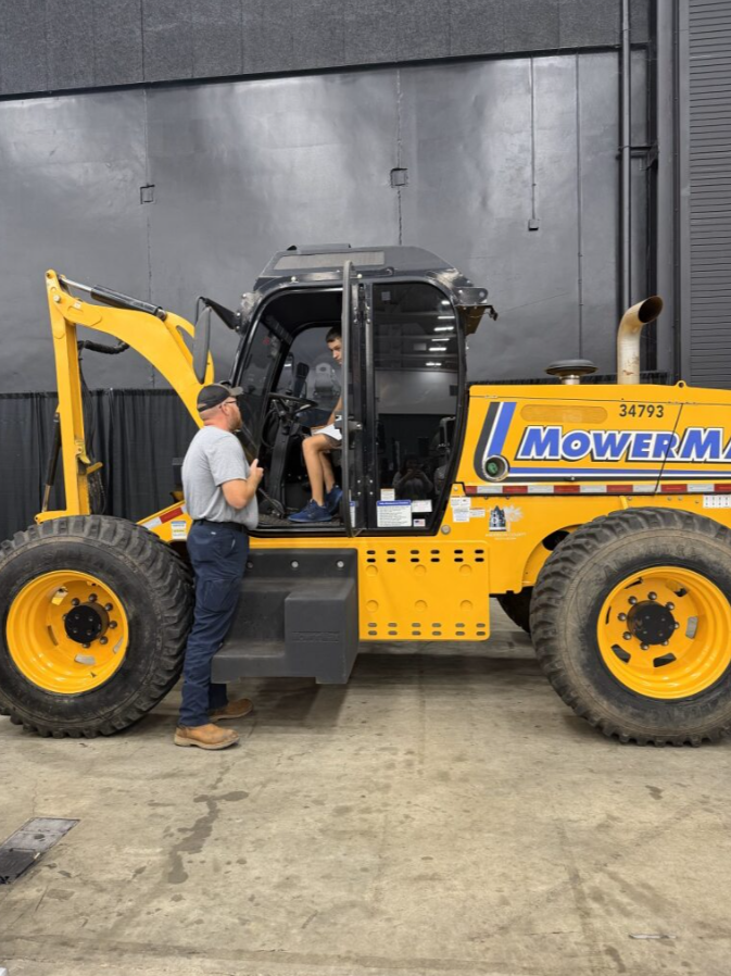 A person in gray shirt and dark pants standing outside a yellow Mower M tractor, talking to a woman sitting inside the vehicle's cab, which has an open door, in an indoor setting with a concrete floor and dark walls.