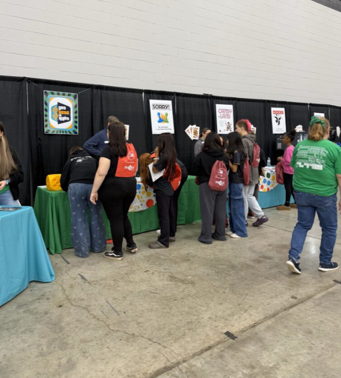 People at a convention or event browsing booths with black backdrop and colorful displays, some wearing backpacks.