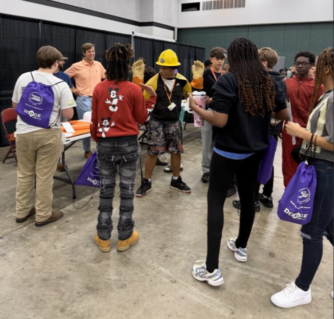 Group of people, including a woman in a yellow hard hat and gloves, gathered around at an indoor event or career fair.
