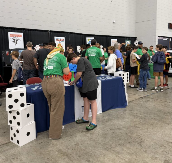 Children and adults at an indoor game and activity fair, with tables featuring large dice and board game posters in the background.