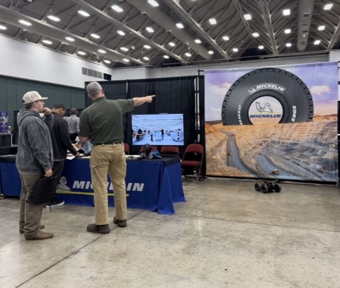People at a Michelin booth in an indoor exhibition, displaying a large tire graphic and a screen showing a racing scene.
