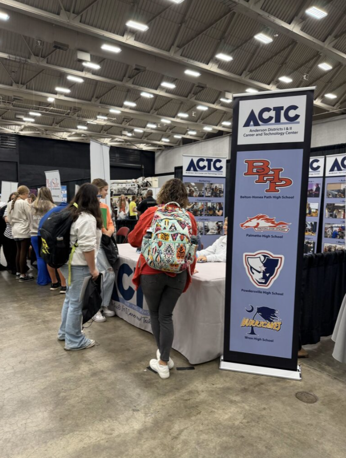 Participants at a career fair booth with banners displaying schools including Benton-Monroe Path High School, Palmetto High School, Powdersville High School, and Wren High School, inside a large indoor venue.