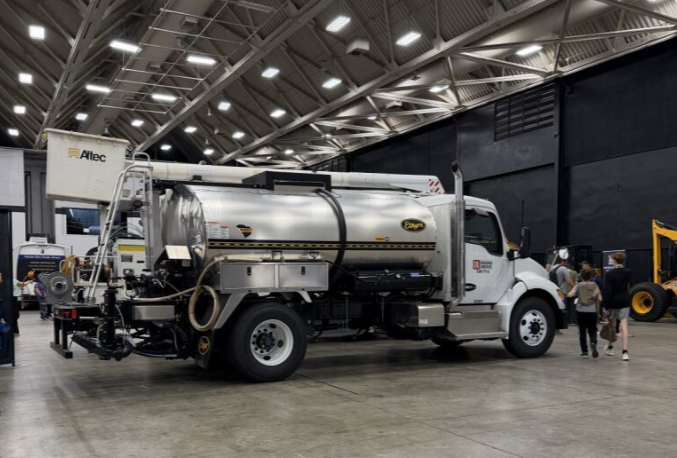 A white vacuum truck with a large metal tank and hoses is displayed indoors at an expo or exhibition, with several people walking nearby.