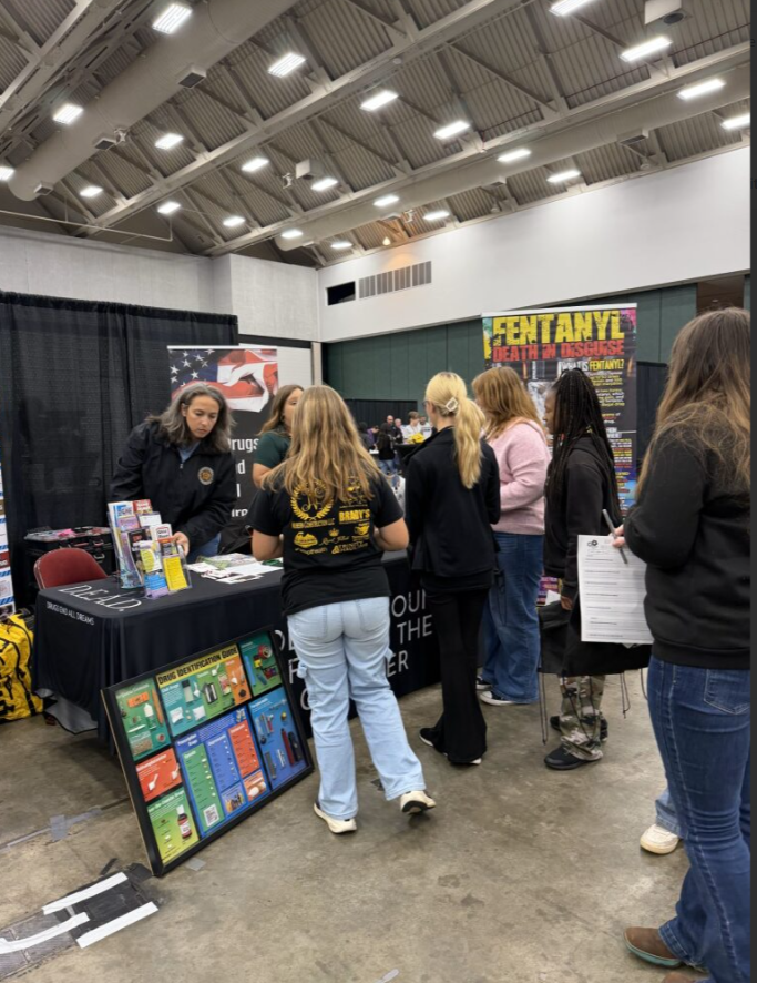 People gathered at an indoor event booth, with informational flyers and displays on the table, and a large colorful banner mentioning 'Fentanyl'.