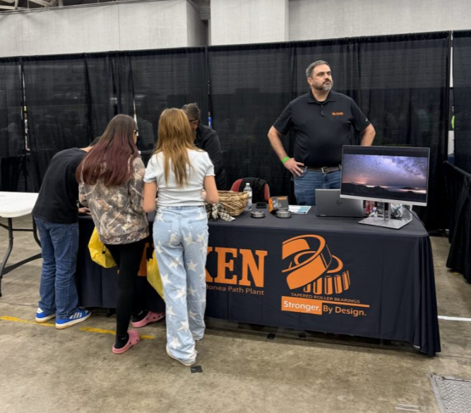 People at a trade show booth with a black tablecloth displaying the logo and name of a company that manufactures roller bearings. The booth has a computer monitor, and the company representative is standing behind the table.